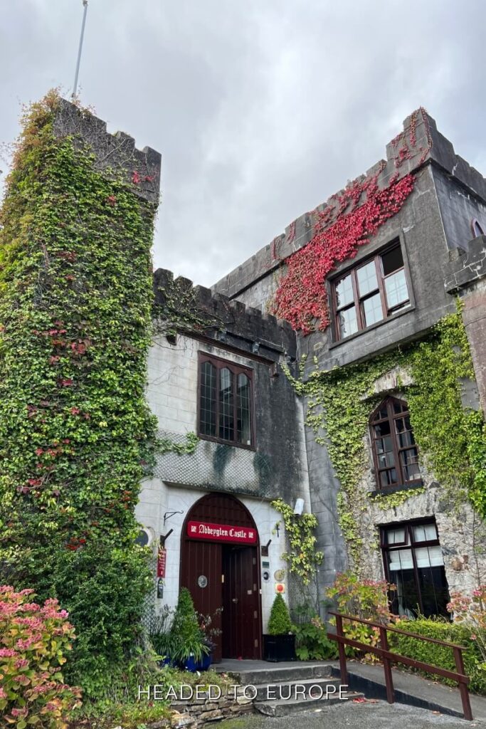 Stone façade of Abbeyglen Castle Hotel, partially covered in red and green ivy, with medieval-style windows and a wooden door framed by plants under an overcast sky.