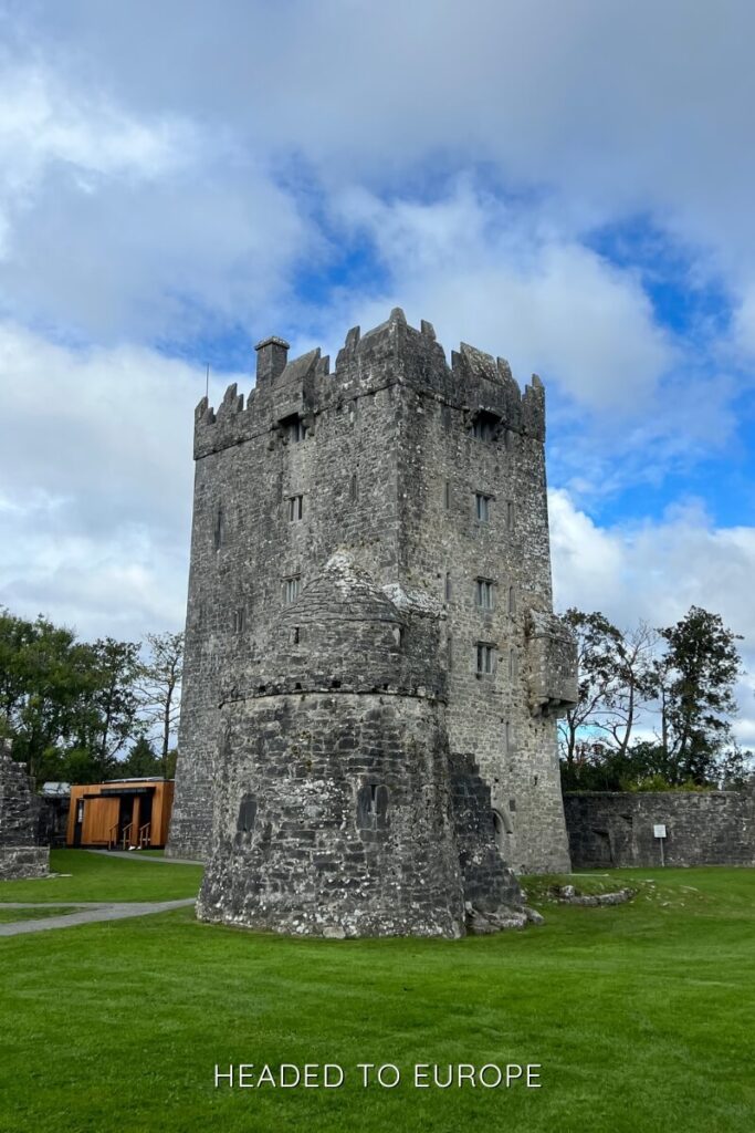 Anghnanure Castle in Ireland stands tall with its grey stone tower, surrounded by green grass and a backdrop of blue sky with scattered clouds.