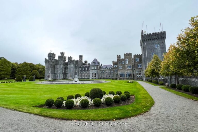 Wide-angle shot of Ashford Castle, showcasing its expansive stone façade, manicured gardens, white flower beds, and central fountain under an overcast sky.