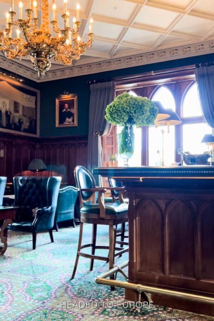 Luxurious castle lounge featuring dark wood paneling, emerald green walls, tufted leather chairs, a grand chandelier, and a vase of vibrant green hydrangeas on a bar counter.