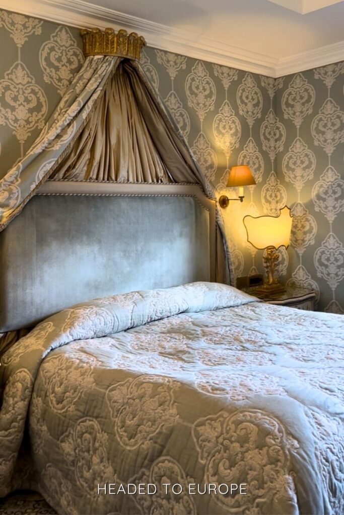 Luxurious Ashford Castle bedroom featuring an upholstered headboard with an ornate canopy crown, damask wallpaper, golden wall sconces, and a lavish embroidered bedspread.