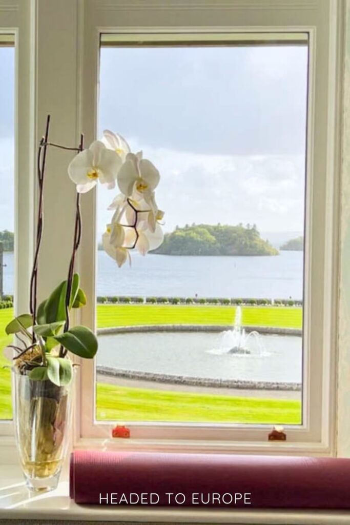 View through a castle window showing a vase with white orchids in the foreground, a circular fountain on a perfectly manicured lawn, and a lake with a small island in the distance under a cloudy sky.