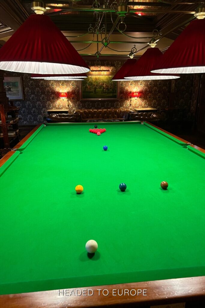 Billiards table set for a game with colorful balls arranged and dramatic red lampshades casting light over the vibrant green surface in a richly decorated room.