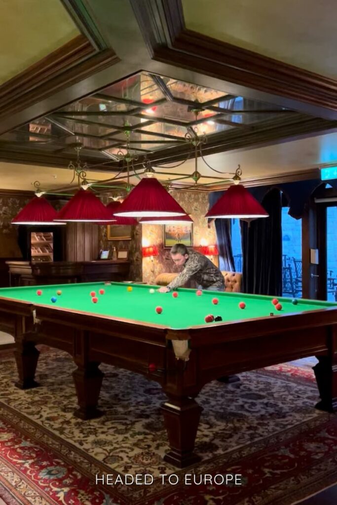Tucker playing snooker on a large green felt table under red-shaded pendant lights in a cozy wood-paneled game room with patterned wallpaper and vintage décor.