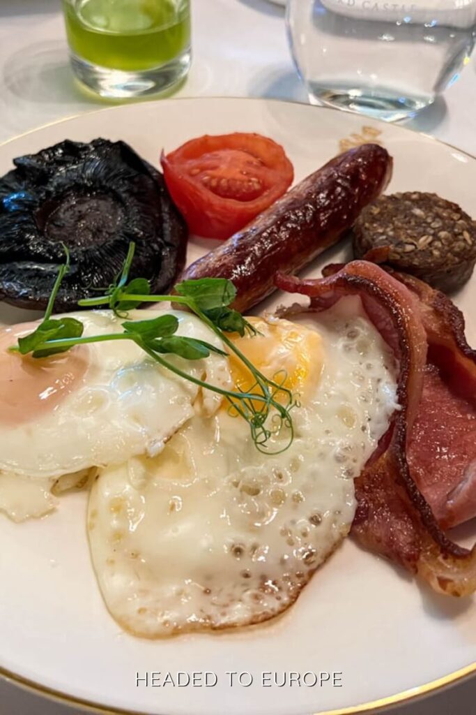 Plate of a traditional Irish breakfast including fried eggs, bacon, sausage, black pudding, grilled tomato, and a large portobello mushroom, garnished with fresh greens.
