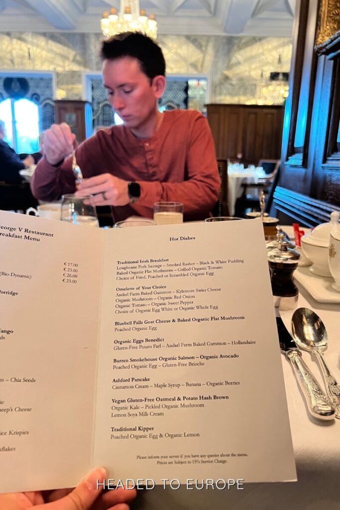 Tucker in a rust-colored shirt stirring his drink at a breakfast table while a menu of hot dishes from the George V Restaurant is held in the foreground.