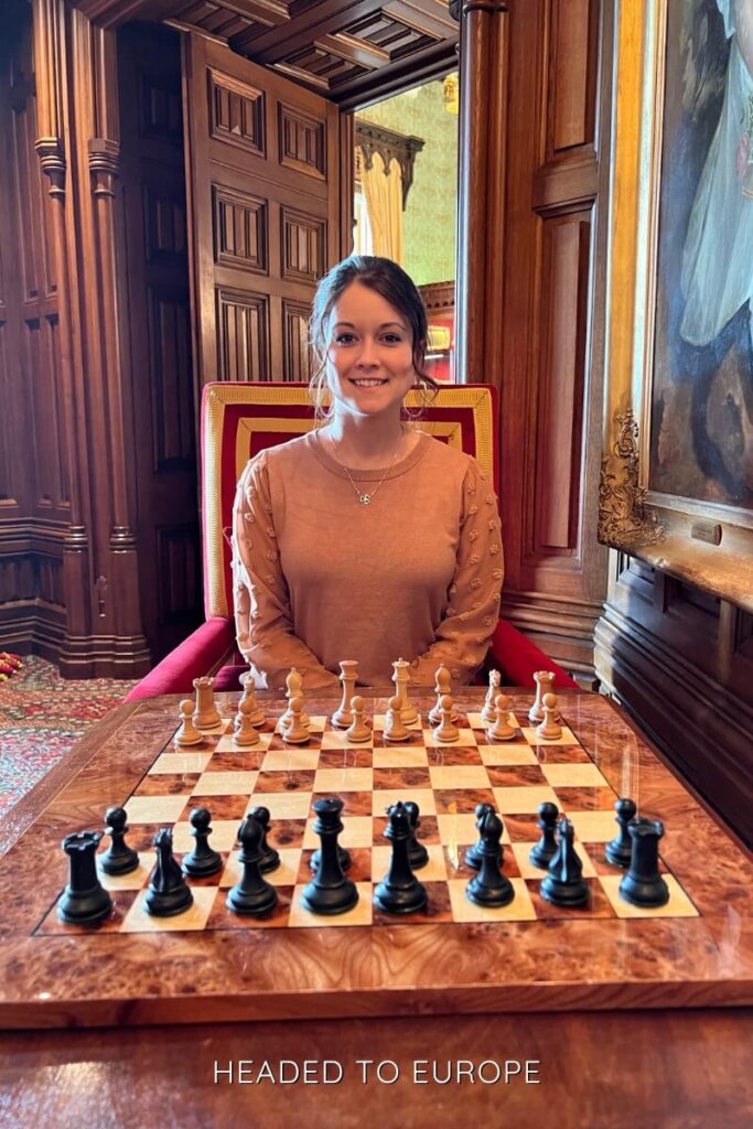 Janae sitting at a chessboard with pieces set for a game, inside a richly paneled wood room with regal decor and a red and gold chair.