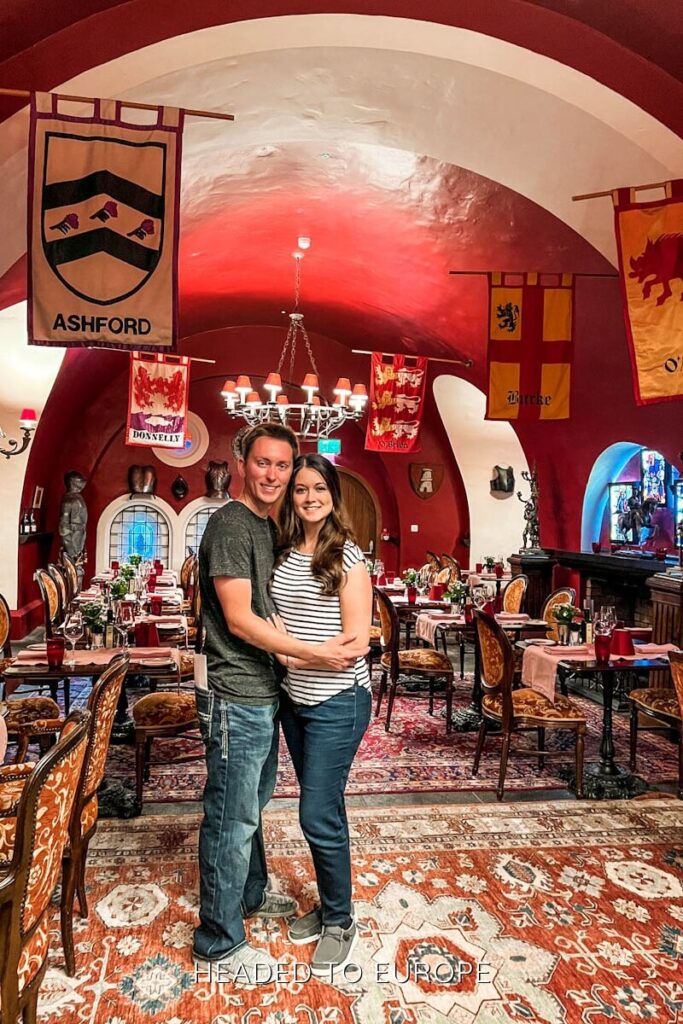 Tucker & Janae embracing in a medieval-style dining dungeon with arched ceilings, heraldic banners labeled “Ashford,” “Donnelly,” and “Burke,” antique furniture, and stained glass windows.