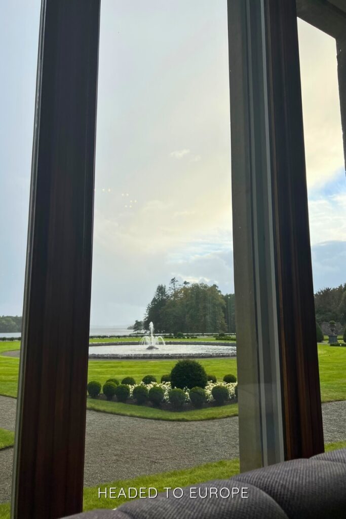 Peaceful view of a manicured garden and a large circular fountain seen through a tall window, with Ashford Castle’s grounds and a lake in the background.