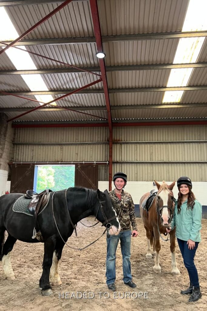 Tucker & Janae standing inside an indoor riding arena next to two saddled horses, wearing helmets and smiling beneath a high metal roof with red beams.