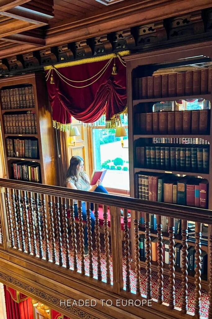 Woman reading a book on a cushioned window seat in an opulent library, surrounded by tall wooden bookshelves, spiral bannisters, and red velvet curtains with gold trim in Ashford Castle.