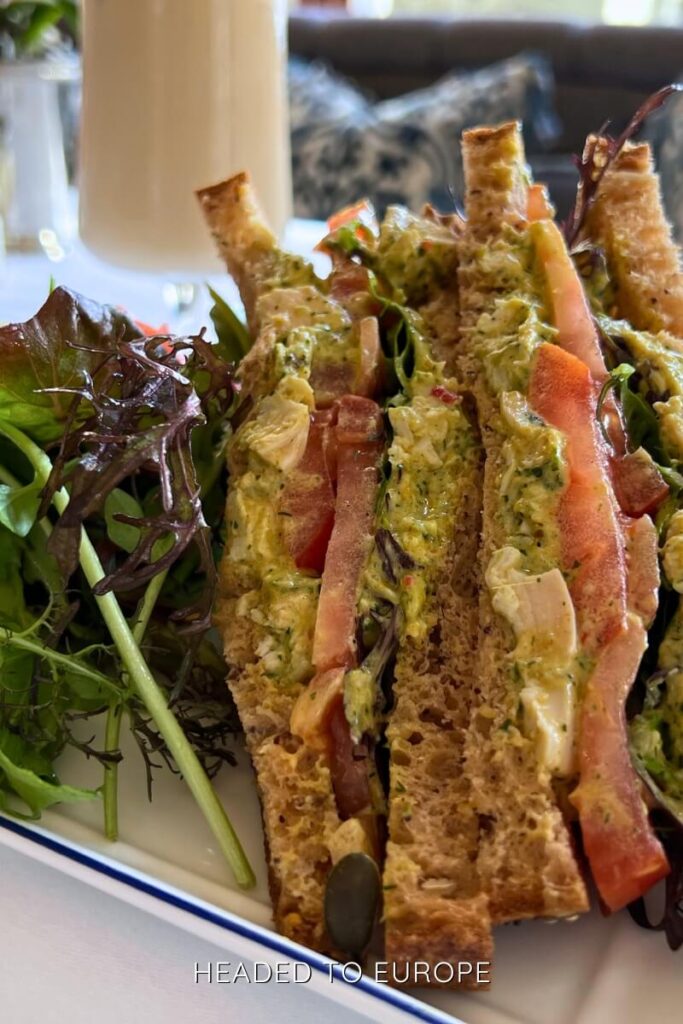 Close-up of a gourmet club sandwich with shredded chicken, tomato, greens, and creamy dressing between slices of brown bread, served alongside a mixed leaf salad.