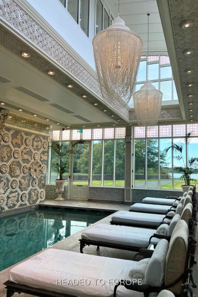 Serene indoor spa area with white cushioned lounge chairs, towering beaded chandeliers, ornate wall carvings, and large windows looking out to a green landscape.