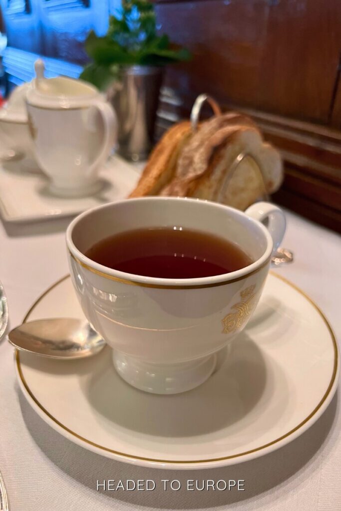 Close-up of a white teacup filled with black tea on a matching saucer, with a silver spoon and toast rack in the background on a formal table setting.