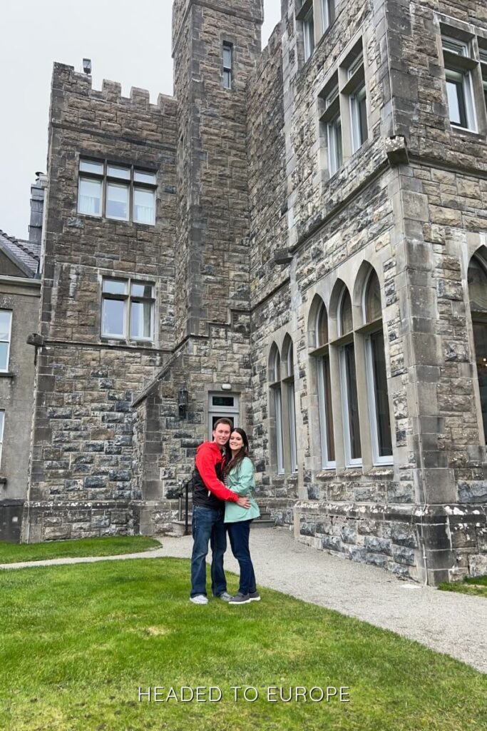 Tucker & Janae hugging on the lawn in front of Ashford Castle’s stone façade, surrounded by medieval-style towers and arched windows on a cloudy day.