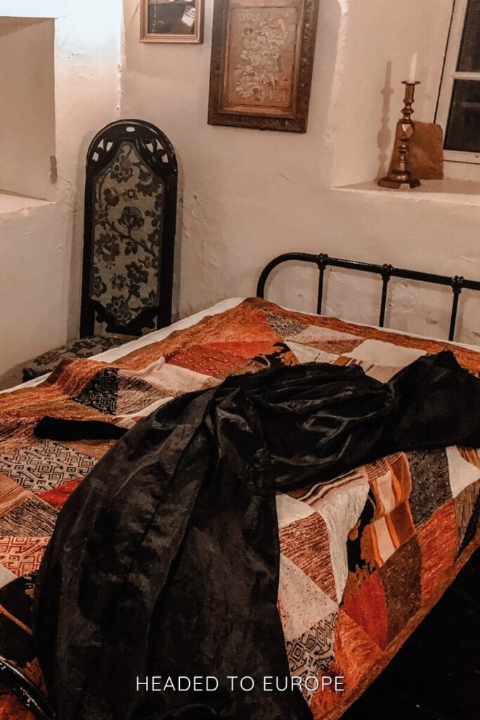 A cozy interior shot of a historic room at Ballygally Castle, showcasing a wrought iron bed with a warm patchwork quilt and a long black cloak draped over it, under soft lighting and vintage decor.