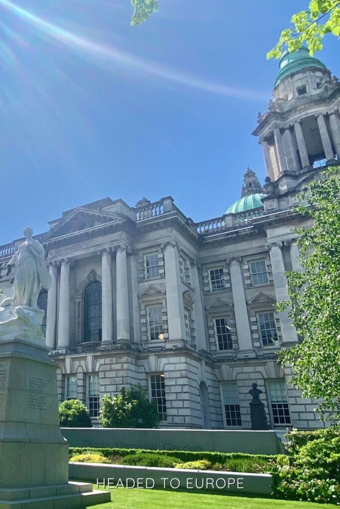 The grand neoclassical facade of Belfast City Hall with its domed green roof, white stone columns, and a statue in the foreground, bathed in sunlight against a clear blue sky.