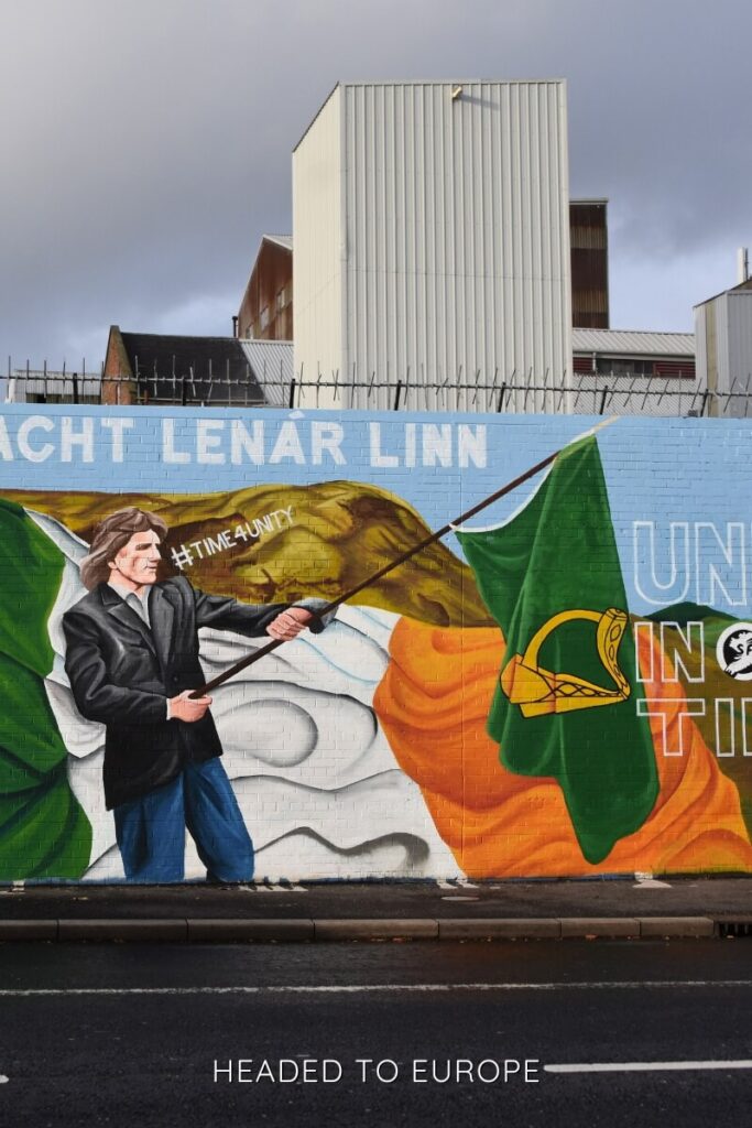 A colorful political mural in Belfast shows a man waving an Irish flag with large Irish tricolor flags in the background and bold lettering on the wall.
