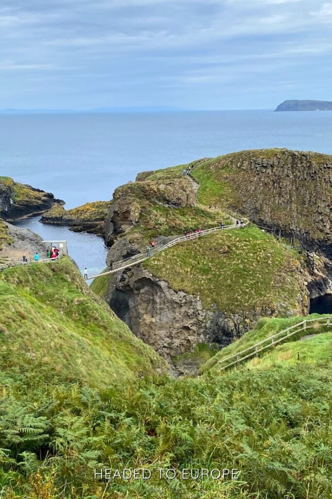 A long rope bridge connects two grassy cliffs over deep blue water near Ballintoy, with tourists walking along the scenic coastal path under a cloudy sky.