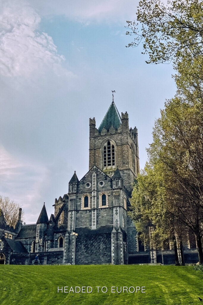 Gothic-style Christ Church Cathedral, one of the most Instagrammable places in Dublin, stands tall with its stone façade and green spire, framed by spring trees and blue sky.