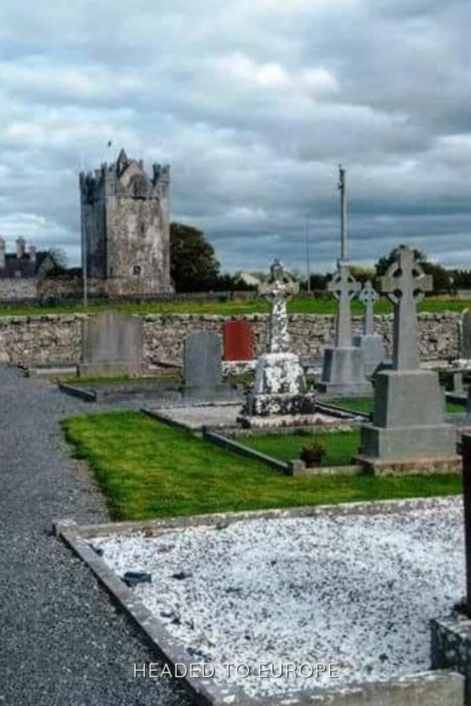 Claregalway Castle rises behind a historic cemetery with traditional Celtic cross headstones and gravel pathways, under a dramatic cloudy sky.