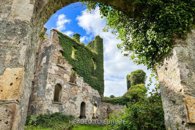 View through a stone archway of the ivy-covered ruins of Clifden Castle in Ireland, with blue skies and scattered clouds above. The castle’s crumbling stone walls and tall greenery-cloaked towers hint at its historic charm.