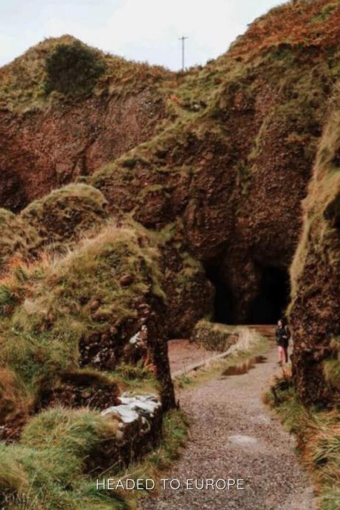 A winding path leads toward a cave opening in the red cliffs of Cushendall, with lush green grass and a hiker approaching the shadowy entrance.