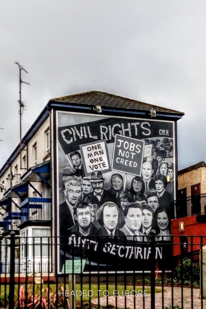 A black-and-white mural in Derry features prominent civil rights figures and signs reading "One Man One Vote" and "Jobs Not Creed," calling for an end to sectarianism.