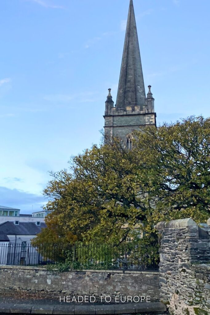 A tall, pointed church spire rises above the trees and rooftops in Derry, set against a light blue sky and framed by a stone wall in the foreground.