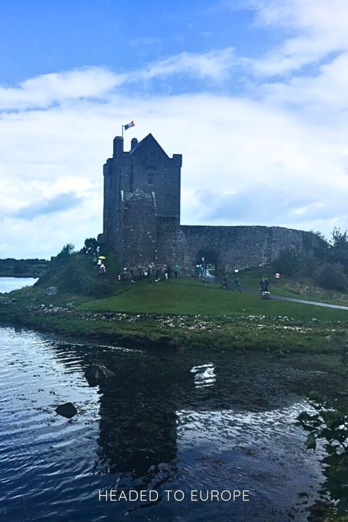 Dunguaire Castle perched on a grassy shore beside reflective water, with tourists walking along a path and the Irish flag flying above the tower.