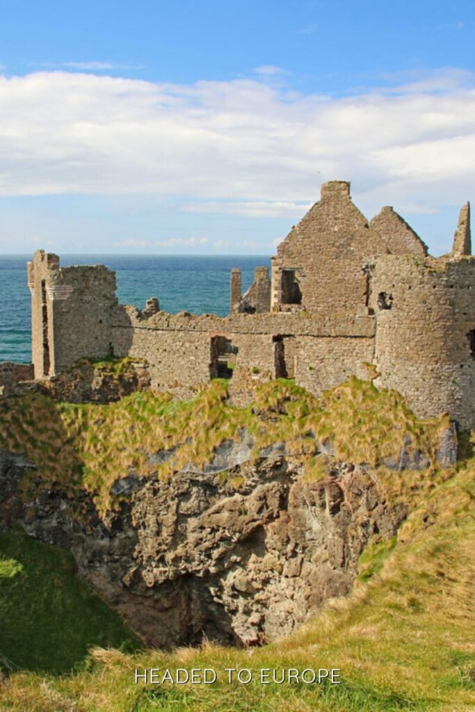 The dramatic cliffside ruins of Dunluce Castle overlook the ocean, with grassy cliffs and ancient stone walls standing against a bright blue sky.