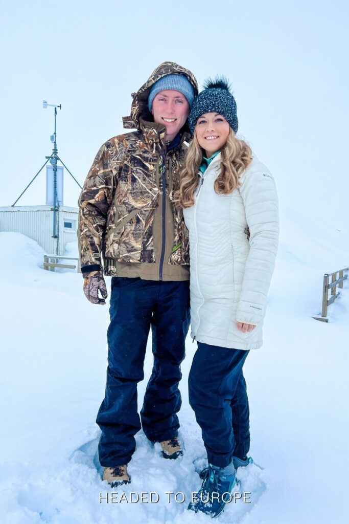 Janae and Tucker smile for a photo at the snowy basecamp of the Into the Glacier tour in Iceland, bundled up in winter gear against the arctic landscape. A weather station and wooden railings are visible behind them in the frozen whiteout conditions.