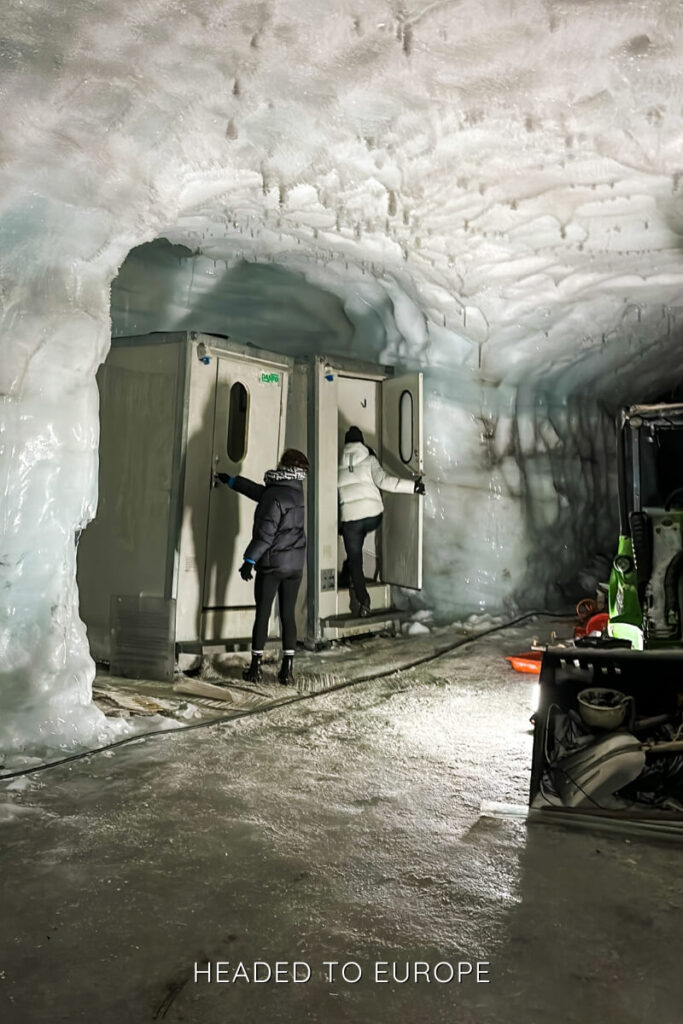 Two women in puffer jackets use restroom facilities built inside the icy walls of the Into the Glacier cave, highlighting the unique infrastructure deep within the glacier. The ice tunnel is dimly lit, with equipment scattered around the rugged frozen floor.