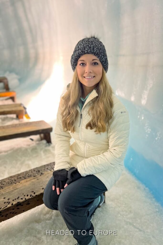 Janae sits on a wooden bench in an icy chapel room inside Into the Glacier, surrounded by glistening blue walls. The setting features several benches on a snow-covered floor, evoking a quiet and serene atmosphere.
