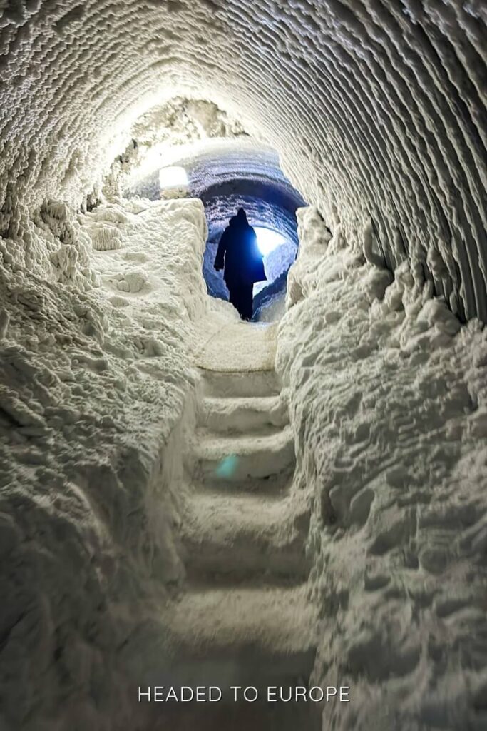A silhouette of a person climbs icy stairs toward daylight at the dramatic arched entrance of the Into the Glacier tunnel. The rough-hewn snow walls frame the glowing exit, giving the scene an ethereal, cave-like atmosphere.