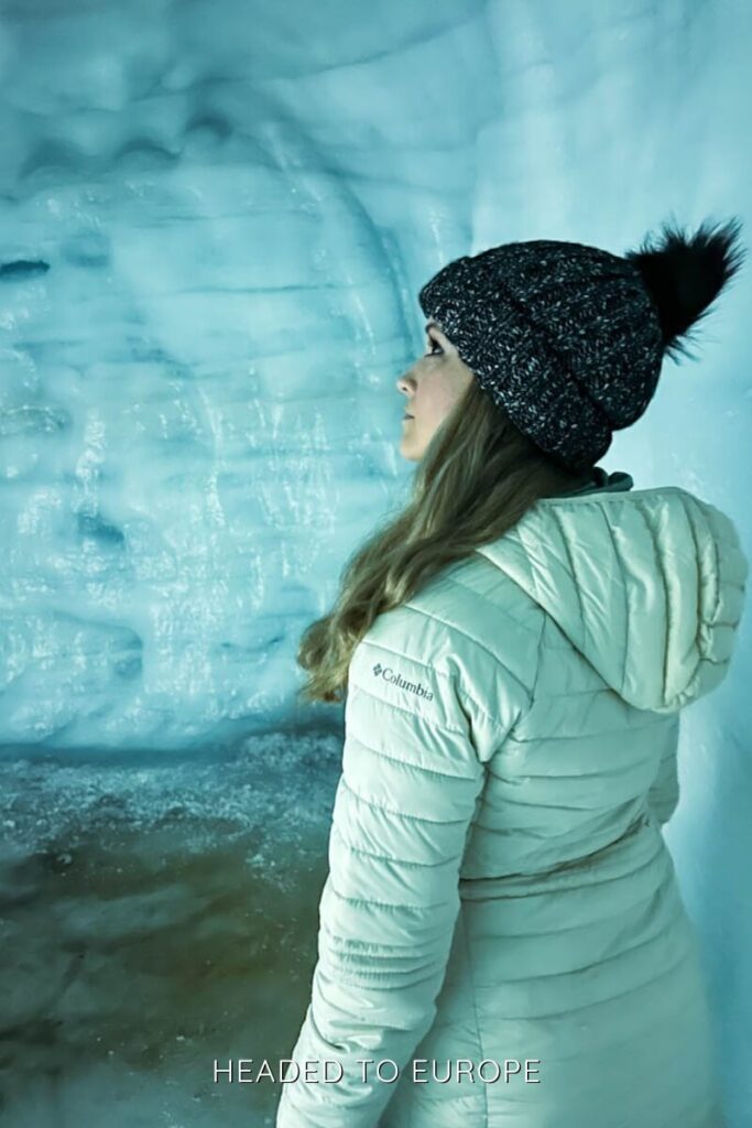 Janae stands inside an ice cave on the Into the Glacier tour, gazing at the glistening walls of the glacier. Dressed warmly in a cream Columbia jacket and knit hat, she appears captivated by the frozen surroundings.