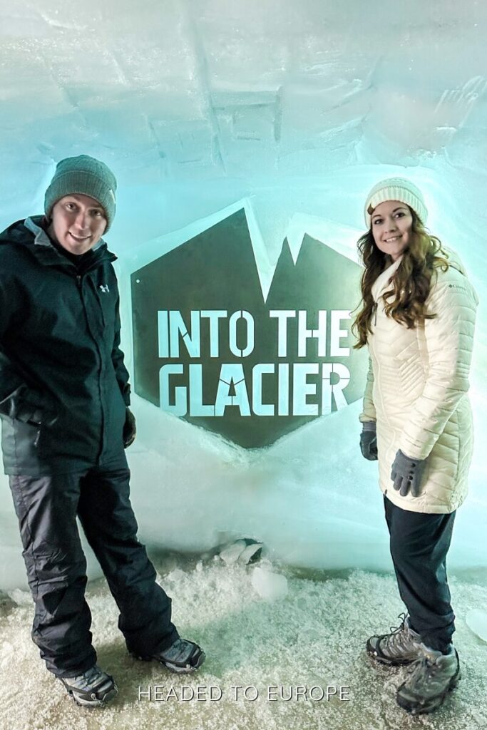Tucker and Janae stand on either side of an illuminated sign reading “Into the Glacier” carved into an icy wall. Both are dressed in winter gear, posing happily at the iconic photo spot that marks the entrance to Iceland’s glacier tunnel experience.