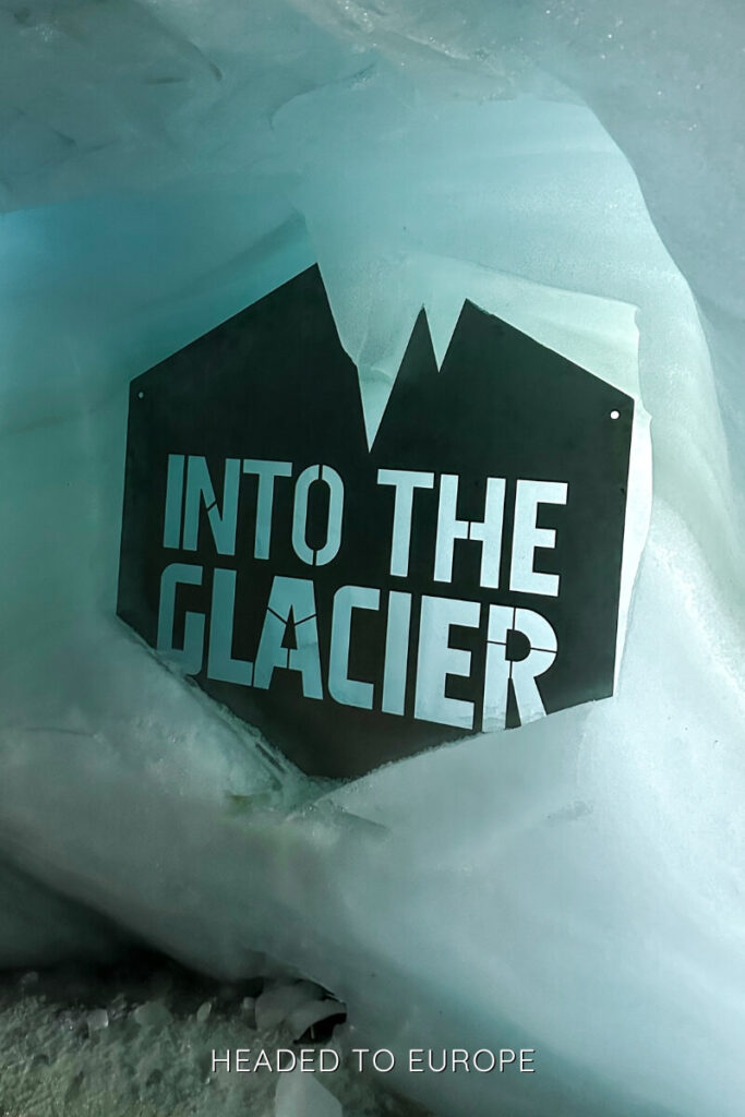 A metal sign reading "Into the Glacier" is mounted on icy blue walls within the glacier tunnel, marking the entrance to the famous Icelandic ice cave tour. The jagged mountain silhouette shape of the sign adds to the adventurous vibe.