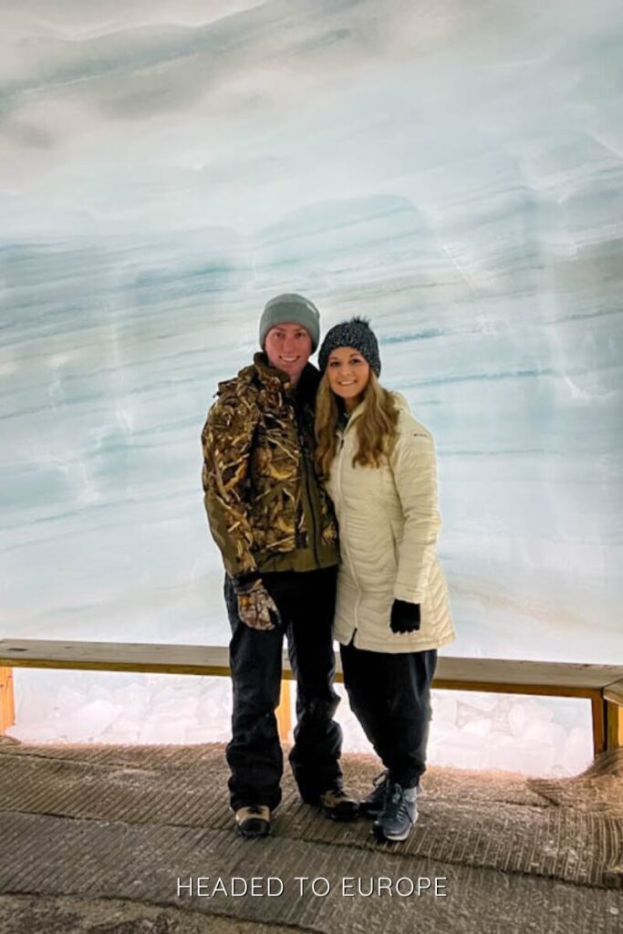Tucker and Janae stand arm in arm inside Into the Glacier, smiling in front of frosty blue glacier walls. A wooden bench behind them contrasts with the icy surroundings of the glacier cave.