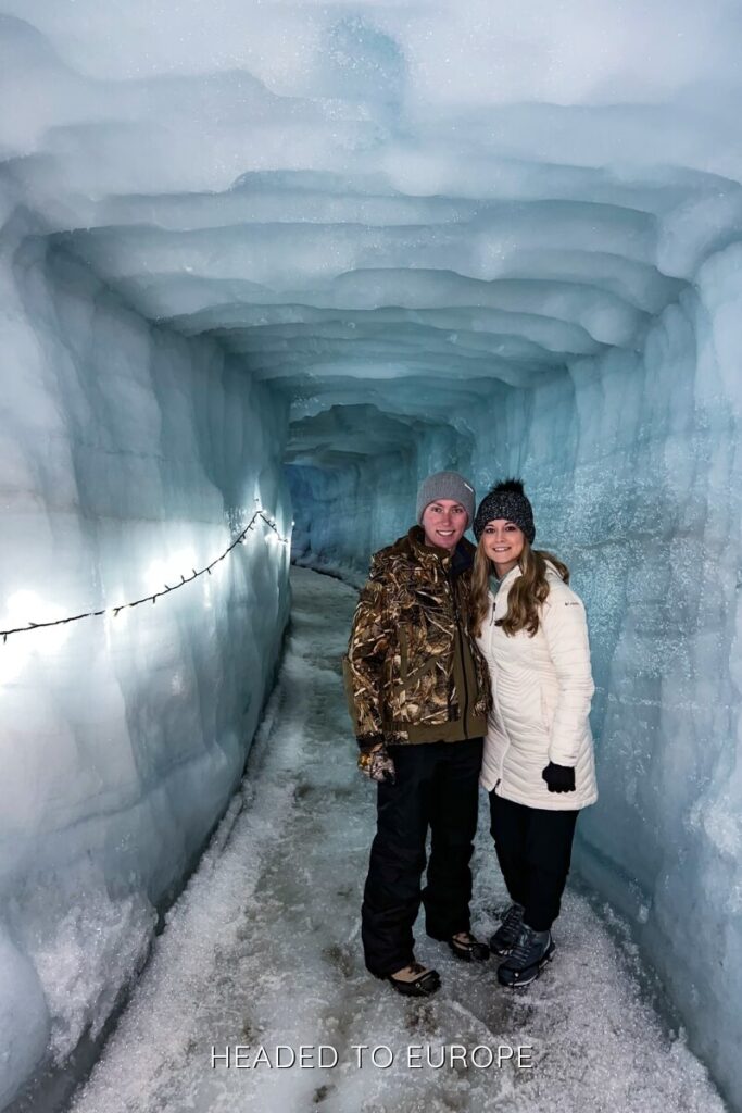 Tucker and Janae stand inside a shimmering tunnel lined with icy walls and string lights during their Into the Glacier tour. The path beneath them is slick and packed, guiding visitors deeper into the glacier.
