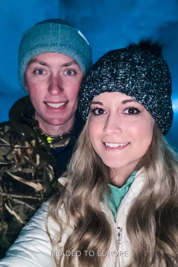 A close-up selfie of Tucker and Janae bundled in warm winter clothes inside Into the Glacier, with the deep blue ice cave walls glowing softly behind them. Their cozy gear and smiles reflect the chilly yet exciting experience.