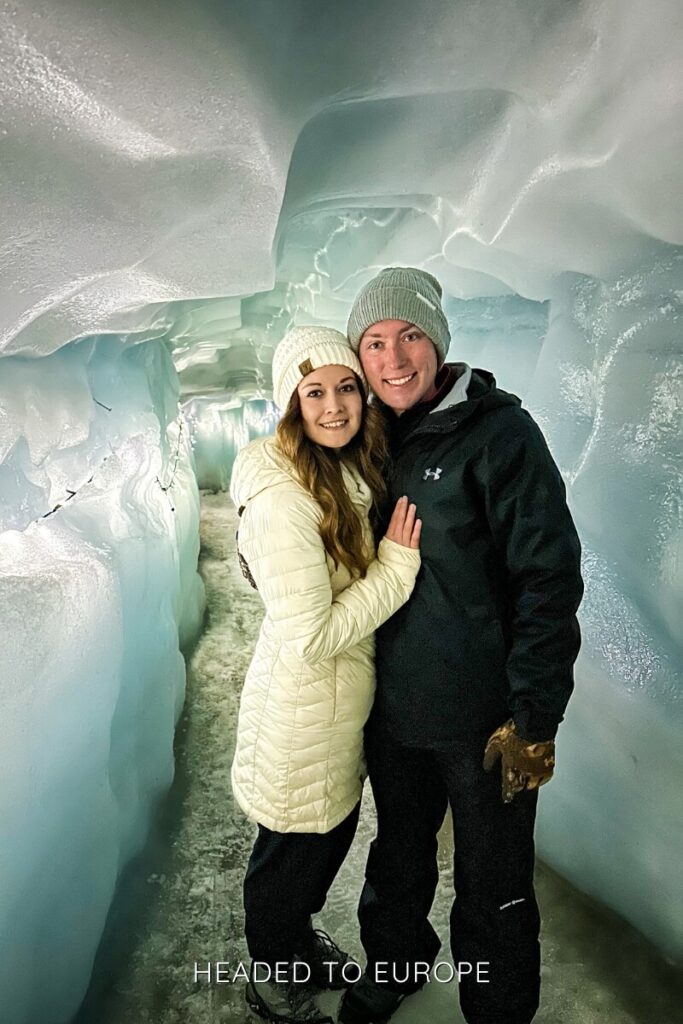 Tucker and Janae smile inside an ethereal tunnel at Into the Glacier, surrounded by curved, pale blue walls of glistening ice. Lights strung along the side highlight the smooth texture of the carved passage.
