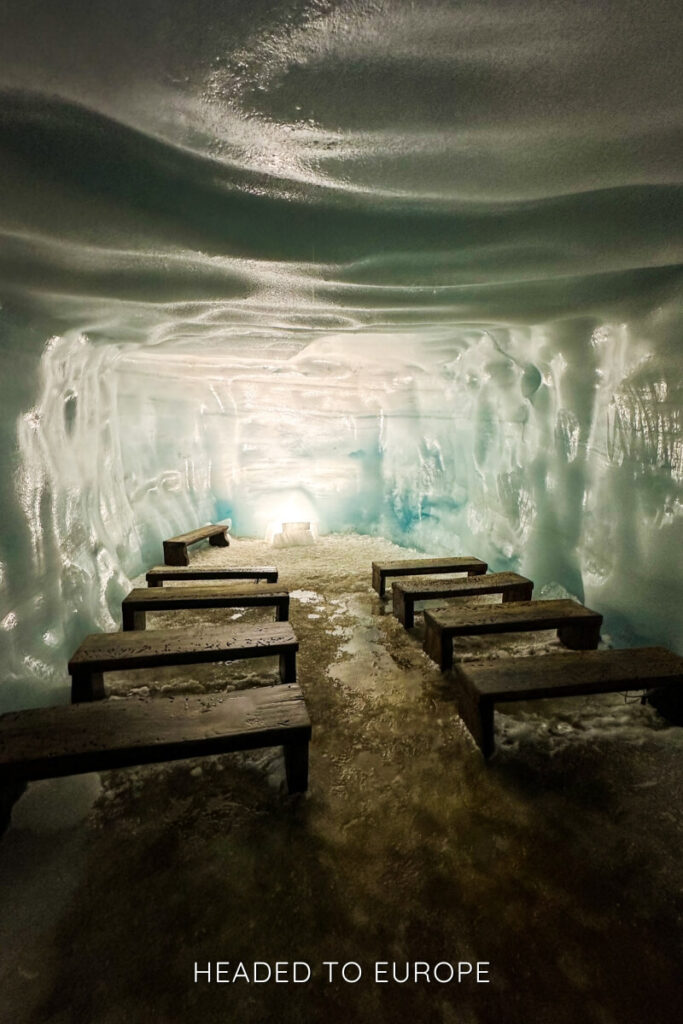 A unique ice chapel inside Into the Glacier, featuring rows of wooden benches on a frozen floor, with softly glowing light illuminating the icy blue chamber. This setting is used for intimate moments and ceremonies deep in the glacier.