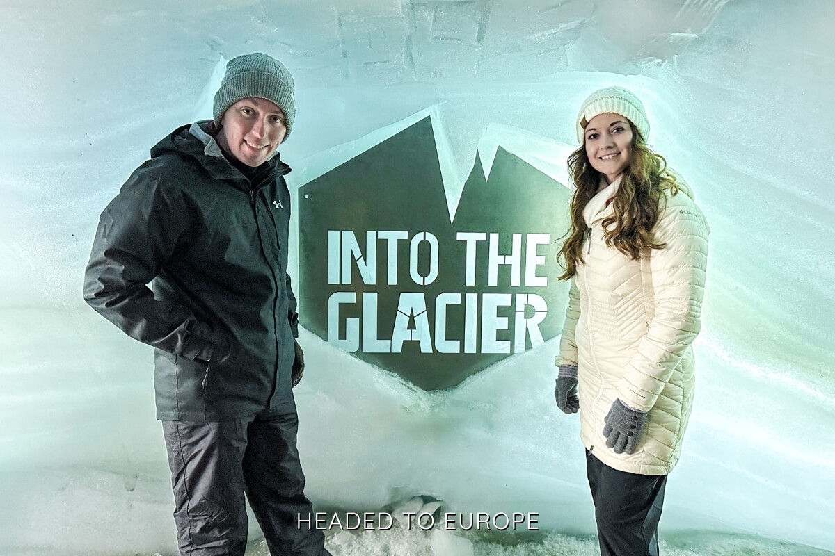 Tucker and Janae pose next to a glowing “Into the Glacier” sign carved into the ice wall, both dressed in light-colored winter coats. This spot is a signature photo opportunity for visitors on the glacier tour.