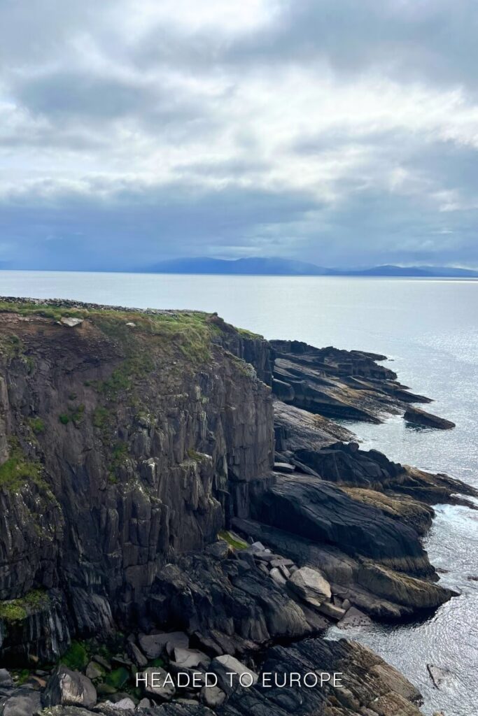 Dramatic rocky cliffs on the coast of Ireland overlooking a calm sea, with layers of jagged stone leading into the ocean beneath a cloudy sky.