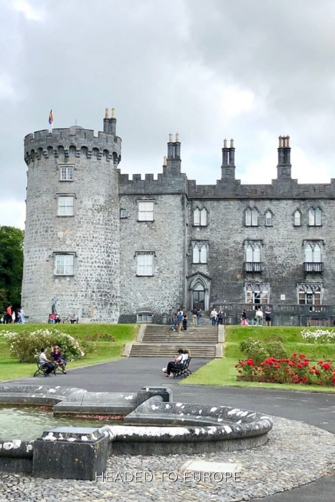 The grand stone Kilkenny Castle with a circular tower and visitors relaxing in the garden, making it a top destination for Weekend Breaks in Ireland.