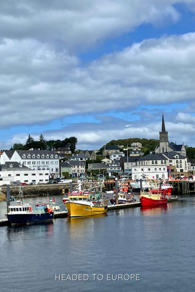 Colorful fishing boats line the waterfront in the harbor town of Killybegs, with a backdrop of small buildings, a church steeple, and cloudy skies.