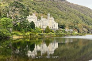 Kylemore Abbey in Ireland reflected in a still lake, surrounded by lush greenery and set against a backdrop of a steep, wooded hillside.