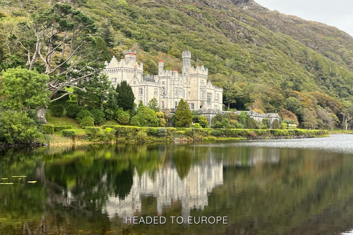 Kylemore Abbey in Ireland reflected in a still lake, surrounded by lush greenery and set against a backdrop of a steep, wooded hillside.