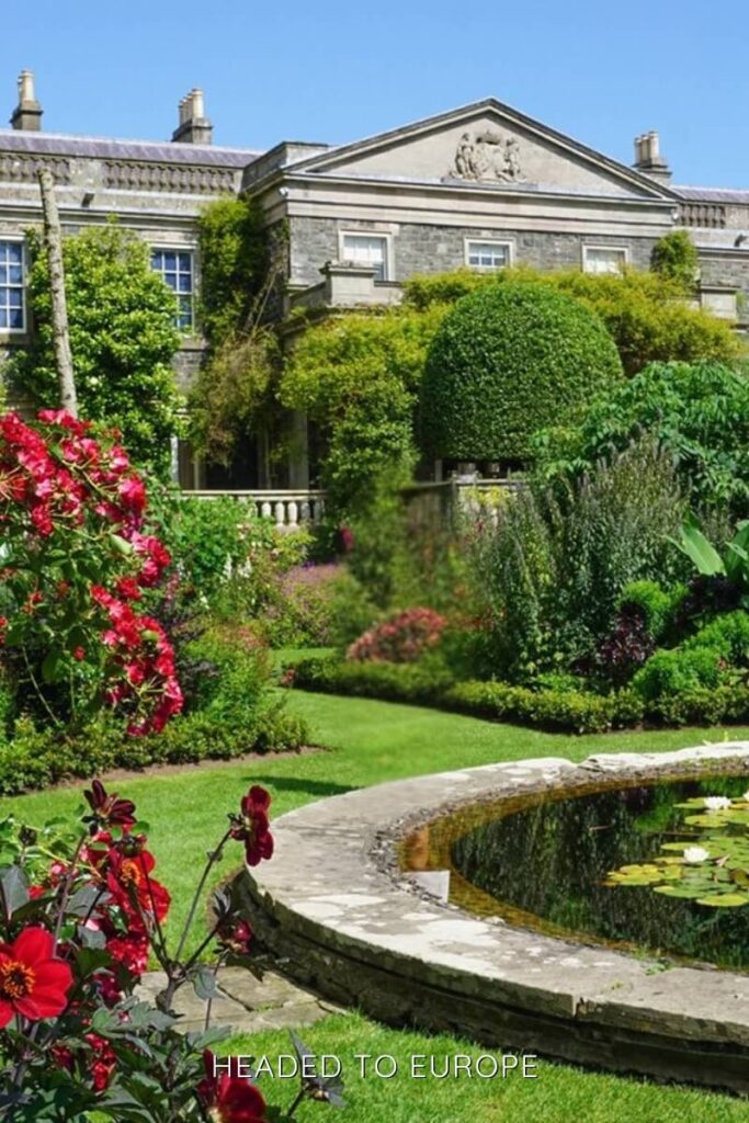 A beautifully landscaped garden at Mount Stewart in Northern Ireland, featuring vibrant red flowers, sculpted greenery, and a circular lily pond in the foreground. The stately stone manor in the background is partially draped in ivy, framed by a bright blue sky.